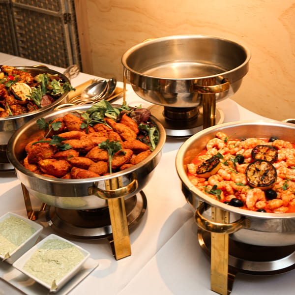 Buffet table with chafing dishes filled with shrimp, falafel, and other dishes, with dipping sauces.