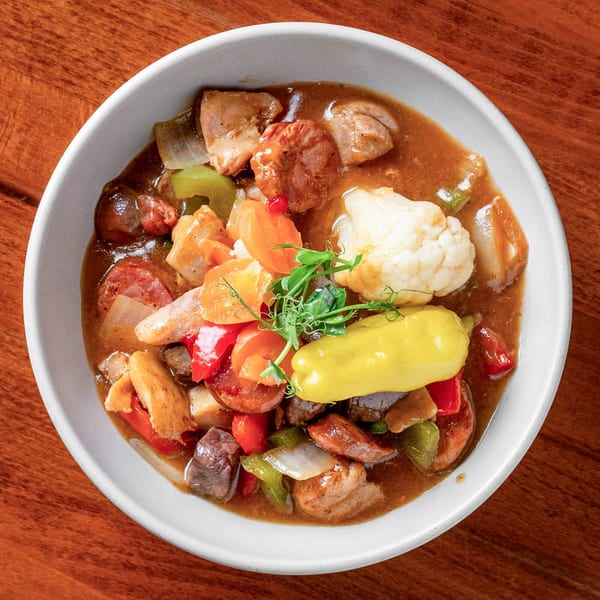 Hearty stew with meat, sausage, cauliflower, peppers, and microgreens in a white bowl on a wooden table.
