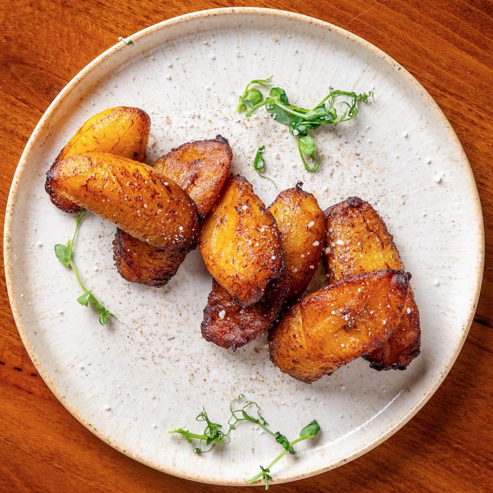 Golden brown fried plantains, sprinkled with salt and garnished with pea shoots on a white plate.