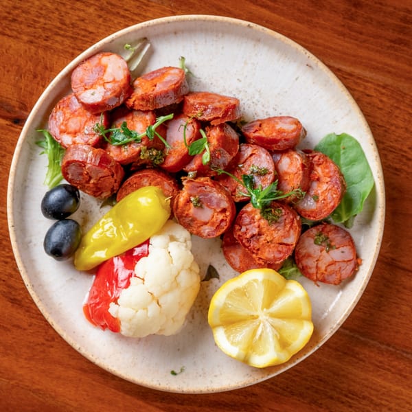 Sliced chorizo sausage with pickled vegetables, olives, and a lemon wedge on a white plate.