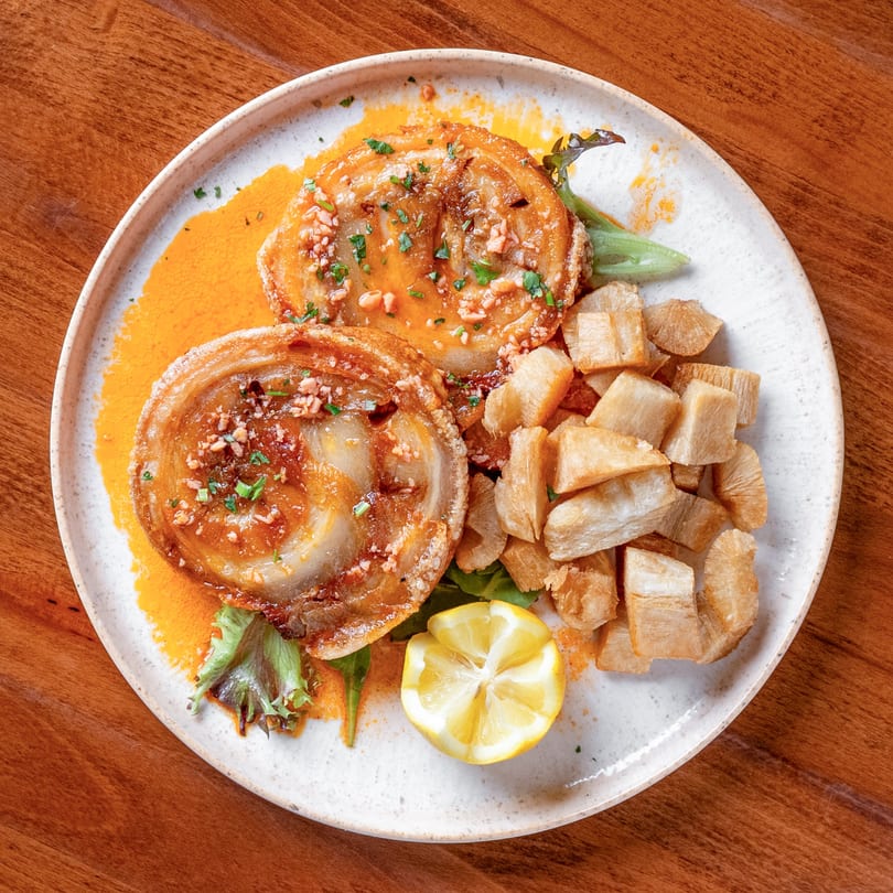 Two crispy pork belly slices with sauce, fried yucca, and a lemon wedge on a white plate.