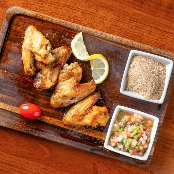 Golden-brown chicken wings on a wooden board with lemon slices, a cherry tomato, and two dipping sauces.