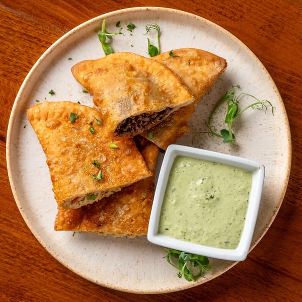 Golden-brown fried pastries with a savory filling, garnished with herbs, served with a side of green dipping sauce.
