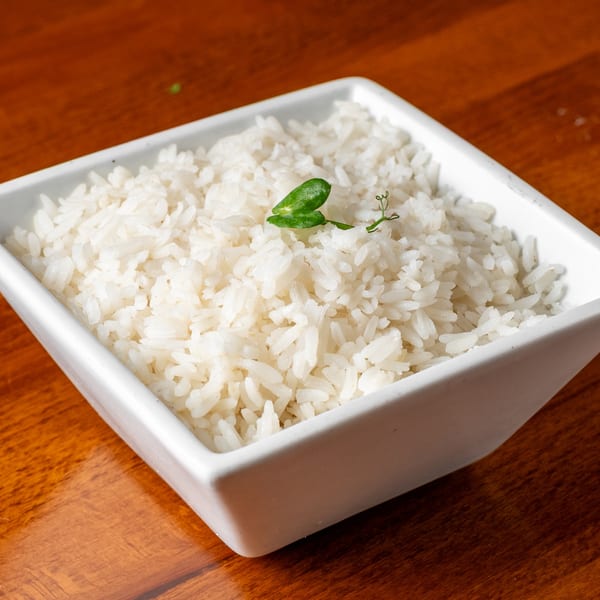 White rice in a square white bowl, garnished with a small green leaf, on a wooden table.