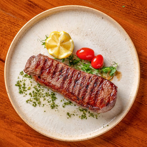 Grilled steak with grill marks, garnished with lemon, cherry tomatoes, and fresh herbs on a white plate.