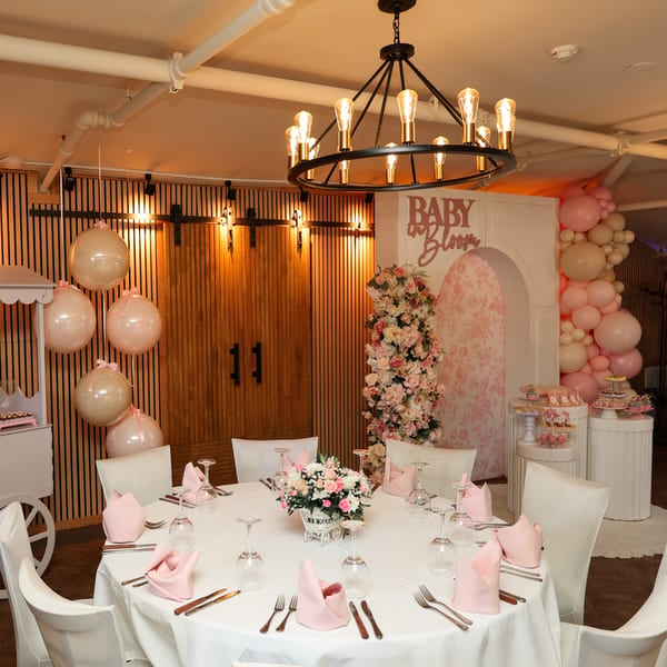 A baby shower setup with a round table, pink and white decor, balloons, and desserts.