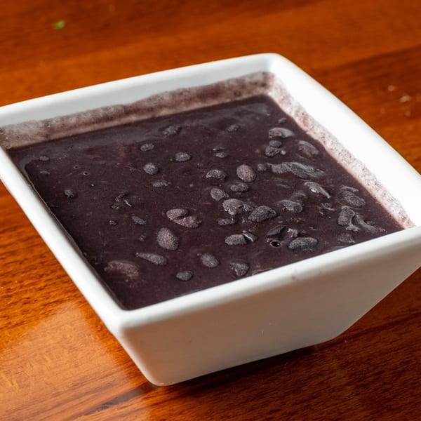 A white square bowl filled with dark, thick black beans on a wooden table.